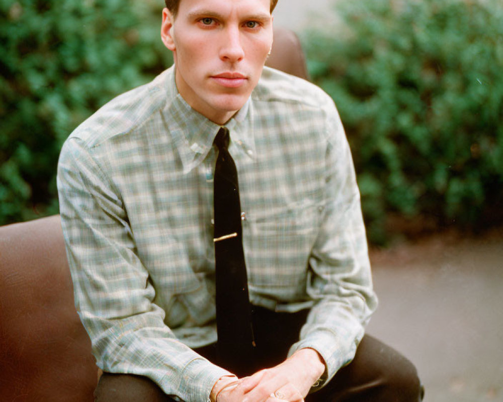 Man in Checkered Shirt and Tie Sitting on Brown Outdoor Couch with Green Shrubbery Background