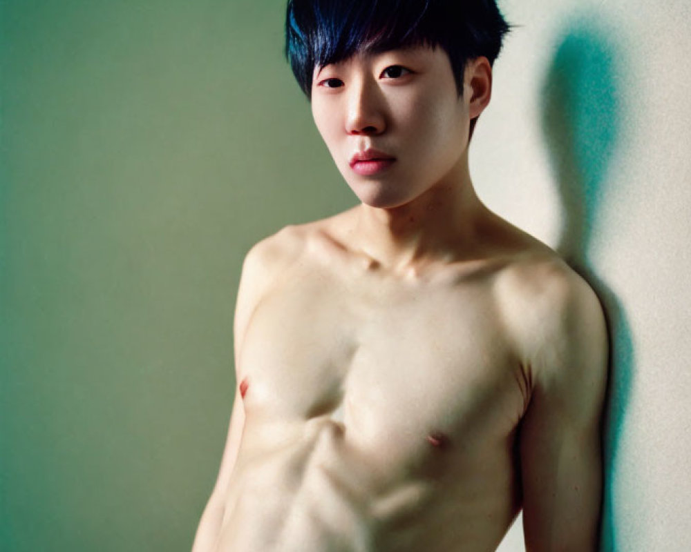 Young man with black hair posing shirtless against pale green background