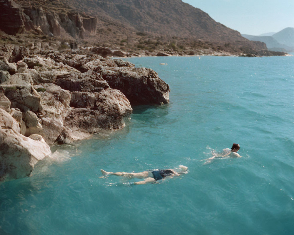 Two people swimming in clear blue waters near rocky shores with hill and sky.