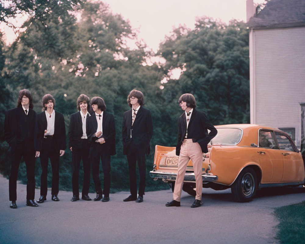 Young men in suits near orange car at dusk with trees and house.