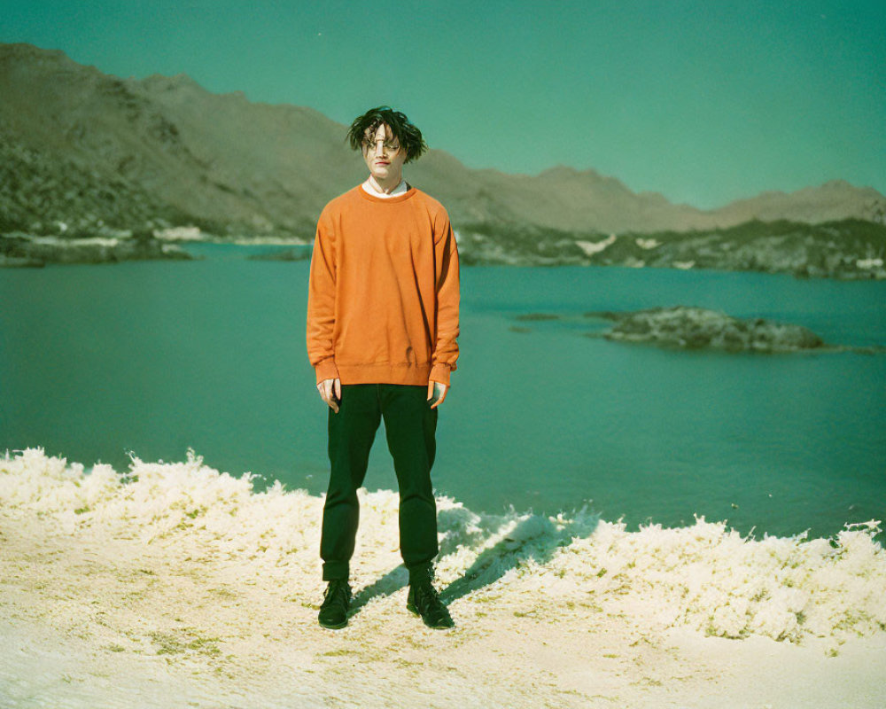 Person in Orange Sweatshirt on Sandy Shore with Lake and Mountain Range