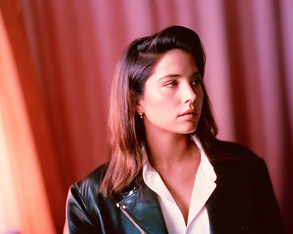 Dark-haired woman in white blouse and leather jacket against pink backdrop with thoughtful expression