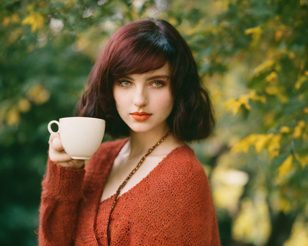 Red-haired woman with nose ring holding cup in red sweater, greenery background