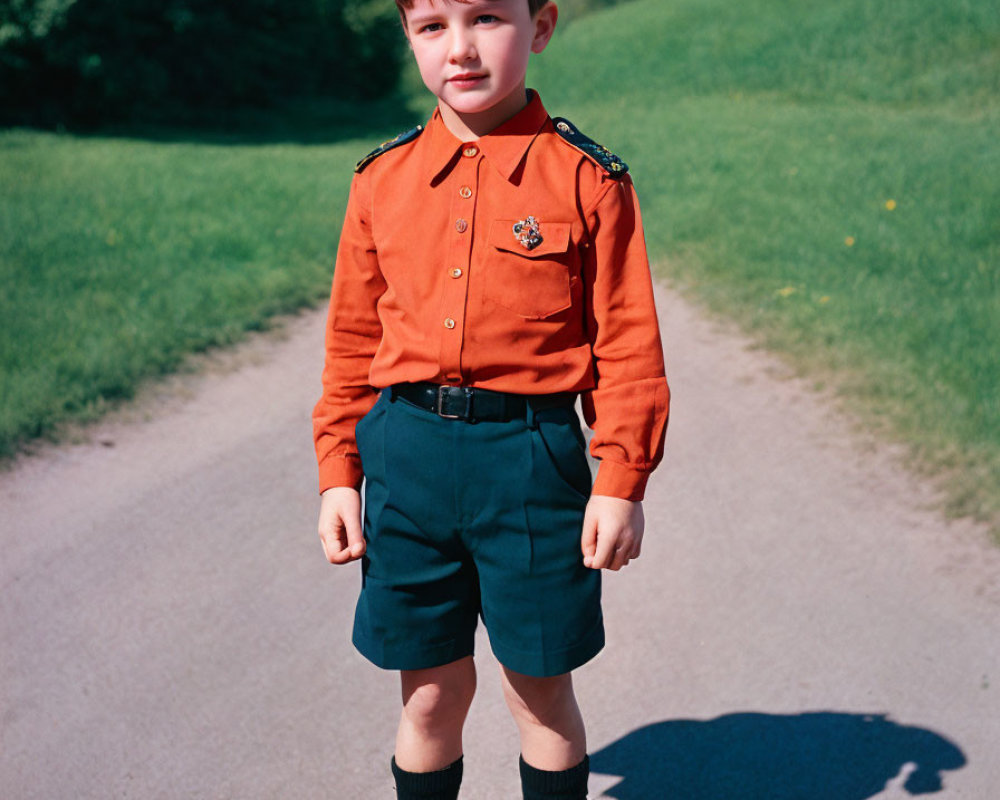 Young boy in orange shirt, black shorts, standing on path with green grass and trees