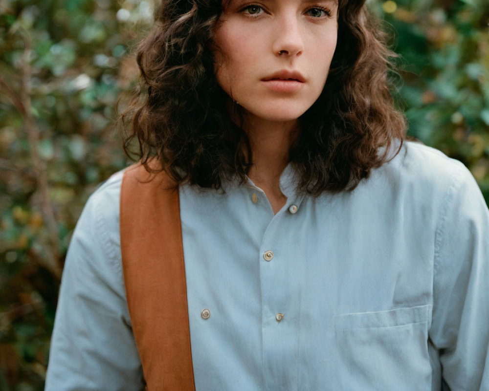 Curly-haired woman in light blue shirt with brown strap, amidst green foliage