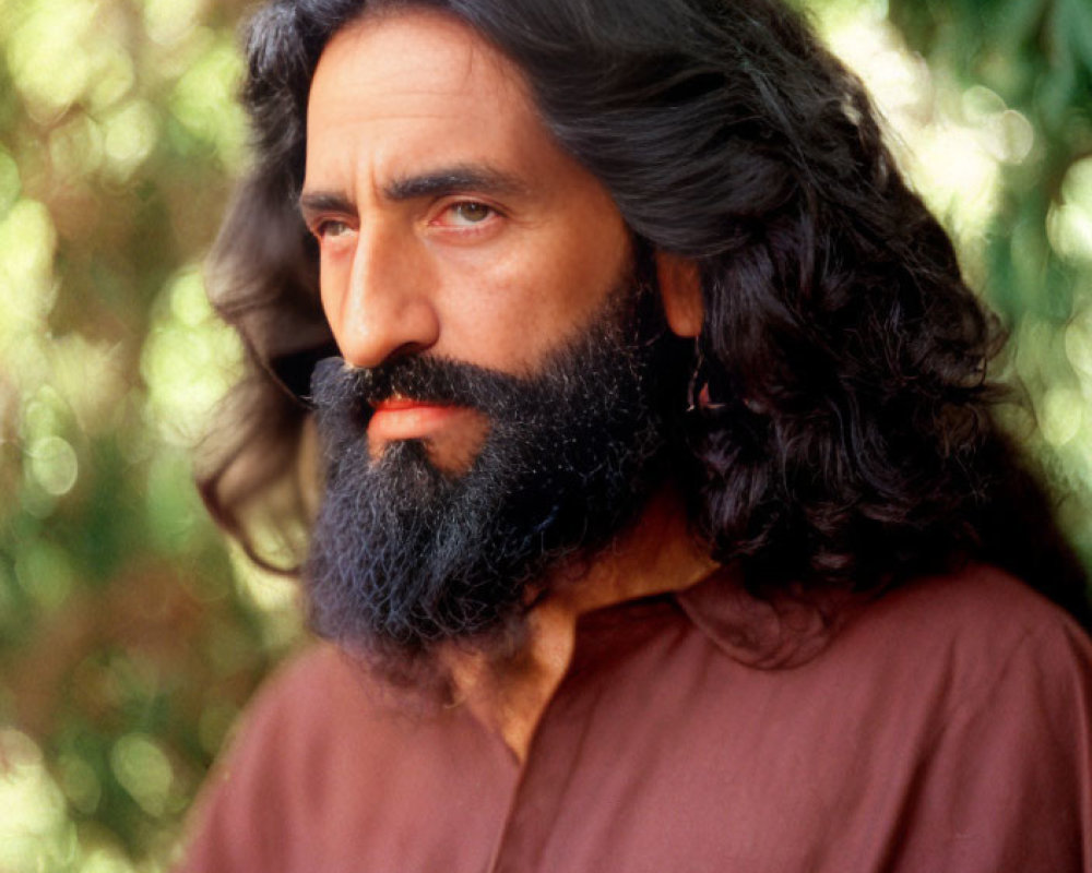 Bearded man with dark hair gazes at camera in front of green foliage