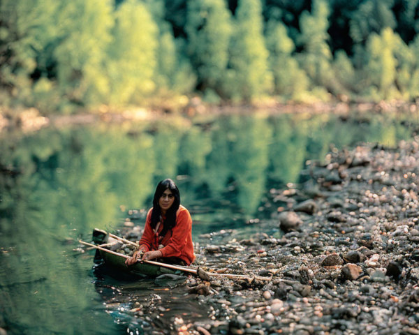 Person in red top on small boat in calm river with greenery & pebbled shore