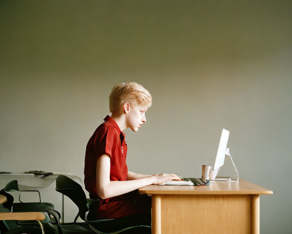 Young individual with short blonde hair at desktop computer in room with muted green wall.